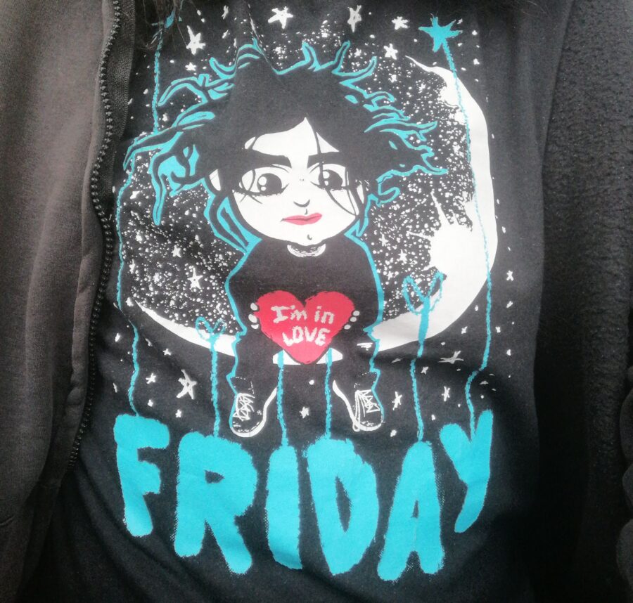Oberkörperselfie, ich trage unter einer schwarzen Hoodiejacke ein schwarzes T-Shirt mit dem Aufdruck "Friday" und einer Zeichnung, die dem Cure-Sänger Robert Smith ähnelt, der auf einer Mondsichel sitzt und ein rotes Herz in Händen mit dem Text "I'm In Love" hält.
