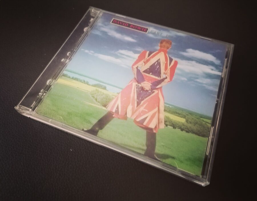 CD "Earthling" von David Bowie. Das Cover zeigt David Bowie in einem Union-Jack-Mantel. Er steht breitbeinig mit dem Rücken zur Kamera, seine Hände halten sich hinter dem Rücken fest. Im Hintergrund ist eine grüne Landschaft mit einem See zu sehen, Am blauen Himmel befinden sich weiße Wolkenfetzen.