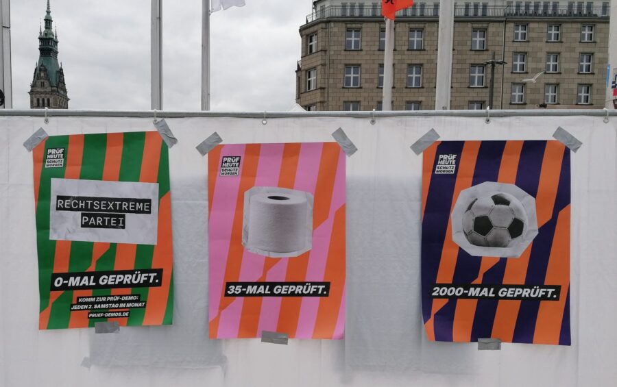 Drei Plakate der PRÜF-Demo: Rechts ein Plakat mit einem Fussball und der Unterschift "2000-mal gepfüft", in der Mitte ein Plakat mit eine Rolle Klopapier und der Unterschrift "35-mal geprüft" und linke ein Plakat mit der Aufschrift "Rechtsextreme Partei" und der Unterschrift "0-mal geprüft".