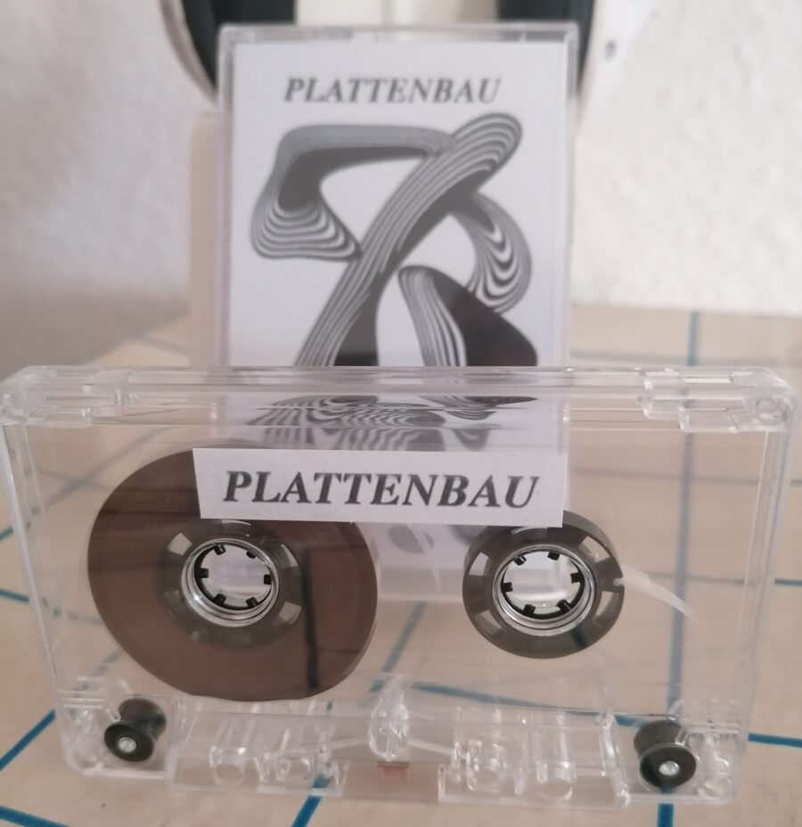 Eine transparente Kassette mit einem Aufkleber "PLATTENBAU" (schwarz auf weiß). Dahinter ist die Kassettenhülle mit Cover zu sehen. Es zeigt eine schwarz-weiße Grafik aus unterschiedlichen Gebilden.