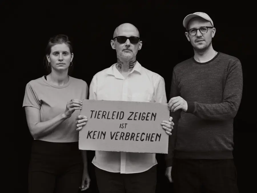 Das Schwarz-Weiß-Foto zeigt die Tierrechtsaktivist:innenAnna Schubert und Hendrik Haßel und in der Mitte den Musiker Moby, der ein Schild in die Kamera hält auf dem "Tierleid zeigen ist kein Verbrechen" geschrieben steht. Er trägt eine Sonnenbrille. Das Foto wurde von Aglaja Brix gemacht.