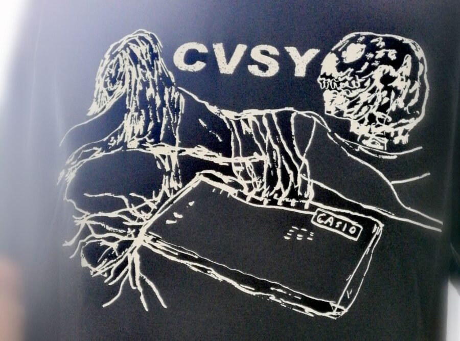 Ein schwarzes T-Shirt mit einem weißen, skizzenartigen Motiv: Oben steht "CVSY". Links sieht man eine Person mit langen, lockigen Haaren, davor liegt ein Gerät mit der Aufschrift "CASIO". Rechts daneben ist eine runde, abstrakte Form, die einem Gesicht ähnelt. Alles ist in klaren, weißen Linien gezeichnet.